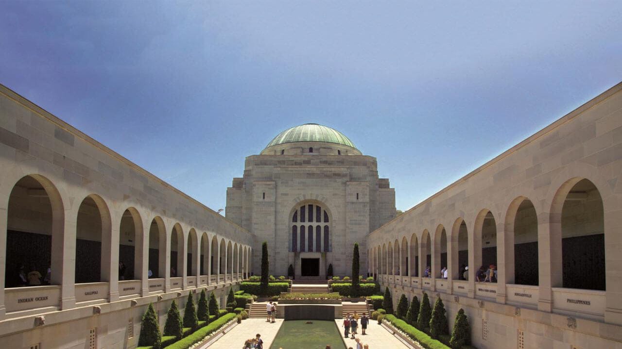 Symmetrical stone memorial with a green-domed central building framed by two long arched colonnades, a rectangular reflecting pool and manicured lawns with visitors on the central walkway.