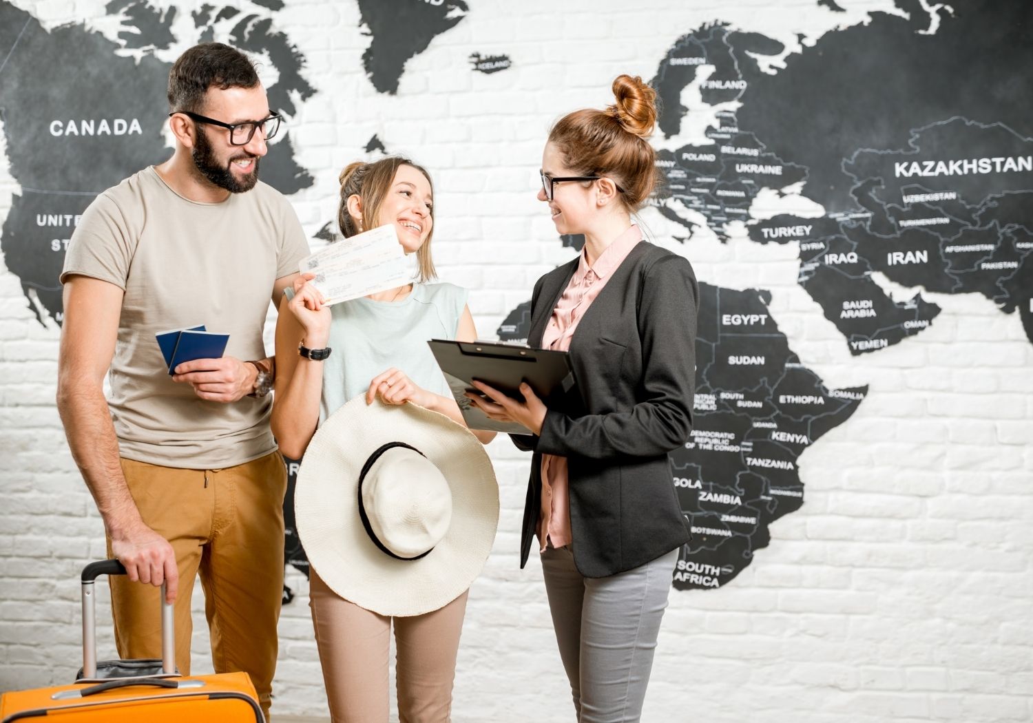 Travel agent RF - Canberra Guided Tours A smiling couple — the woman holding a wide-brimmed sun hat and boarding passes, the man holding passports and an orange suitcase — speak with a travel agent holding a clipboard in front of a large black-and-white world map painted on a brick wall.