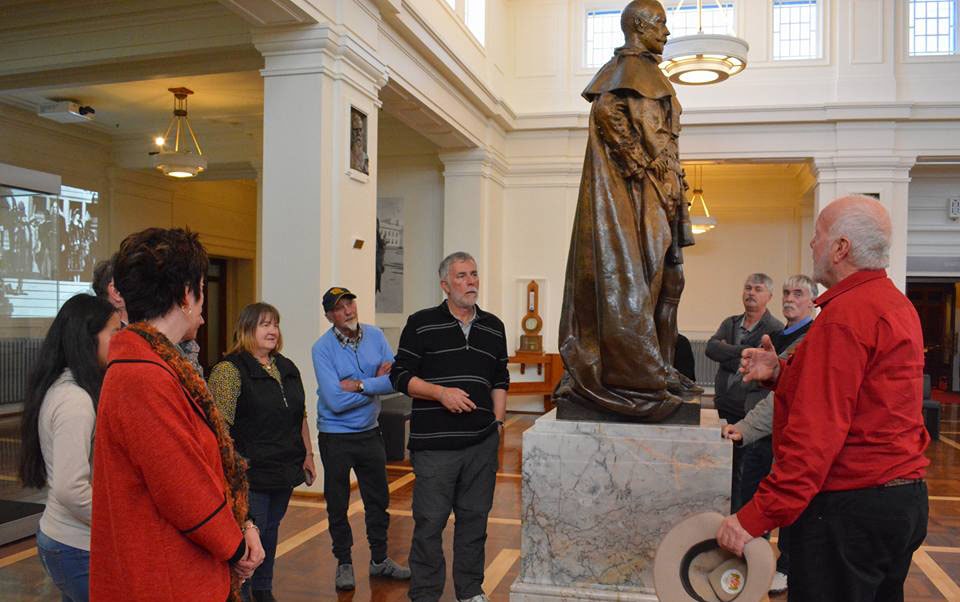 fullday private tour - Canberra Guided Tours A group of visitors stand in a museum gallery around a tall bronze robed statue on a marble plinth as a man in a red shirt gestures while holding a hat, with high ceilings and classical columns in the background.