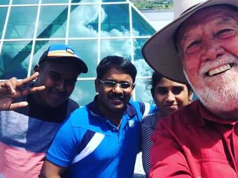 General Neil with Tourists - Canberra Guided Tours Four people smiling for a selfie in front of a glass‑paneled building: an older bearded man wearing a wide‑brimmed hat and red shirt at right, a man in a blue polo and glasses center, a person in a striped top behind him, and another man at left flashing a peace sign and wearing a cap.