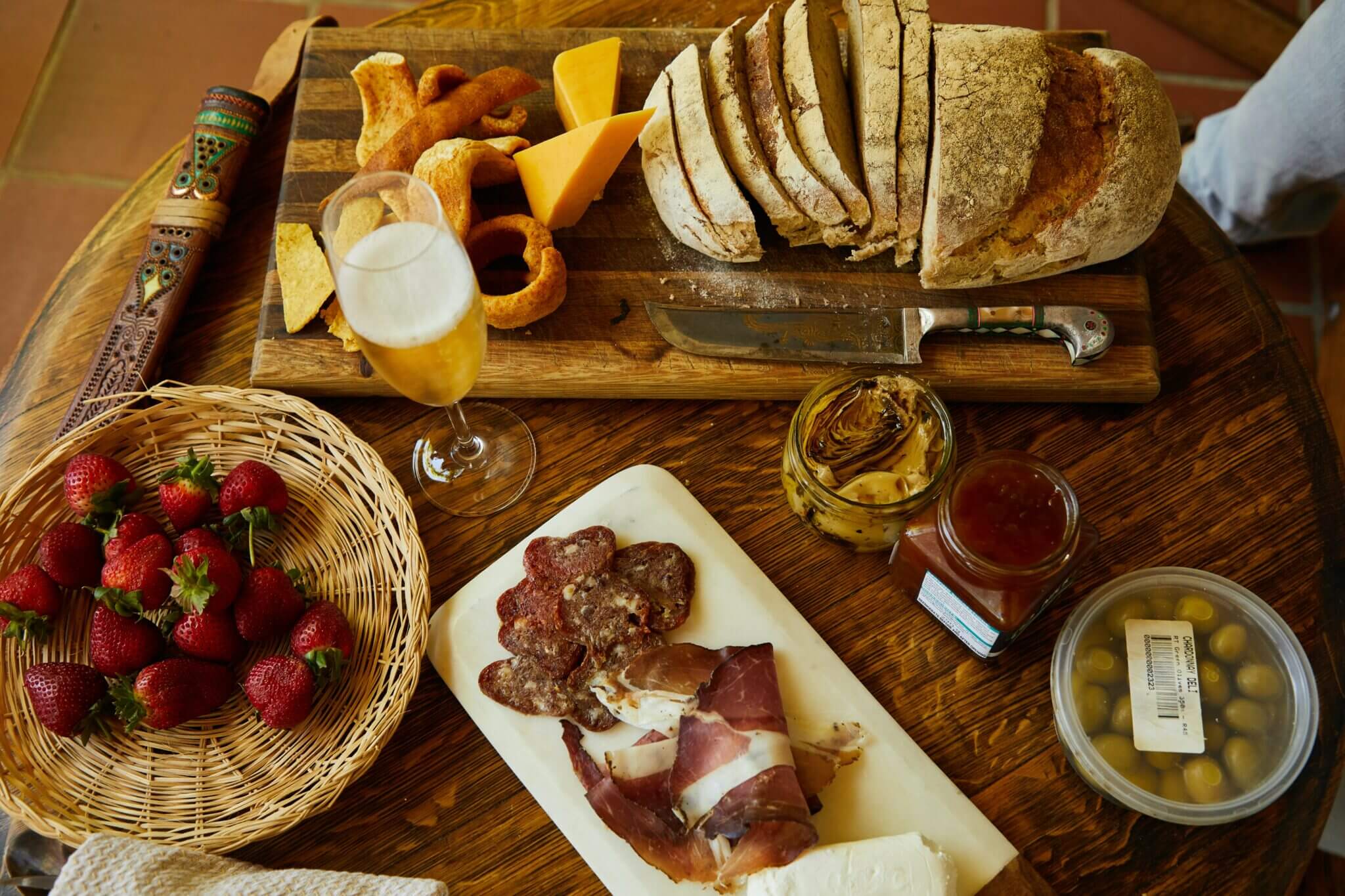 Rustic wooden table with a charcuterie spread: a sliced crusty loaf and knife on a board, cheddar wedges and crackers, a platter of cured meats and prosciutto with cheese, a wicker basket of strawberries, jars of condiments and marinated olives, and a tall glass of beer.