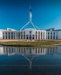 Parliament House in Canberra with its distinctive central flag mast and white colonnaded facade reflected in a foreground pool under a clear blue sky, with a few people on the forecourt.