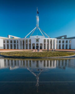 step guide img 01 - Canberra Guided Tours Parliament House in Canberra with its distinctive central flag mast and white colonnaded facade reflected in a foreground pool under a clear blue sky, with a few people on the forecourt.