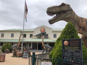 Entrance to the National Dinosaur Museum showing a large lifelike Tyrannosaurus rex head sculpture looming over the courtyard, two long-neck dinosaur models near the museum building and a signboard advertising the museum.