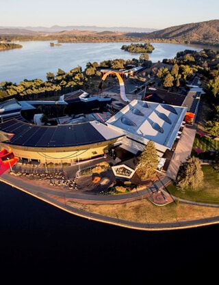 step guide img 03 - Canberra Guided Tours Aerial view of a modern lakeside entertainment complex with curved and peaked white roofs, a waterfront promenade and road at the water’s edge, surrounding trees and a lake stretching to tree-lined hills in the distance.