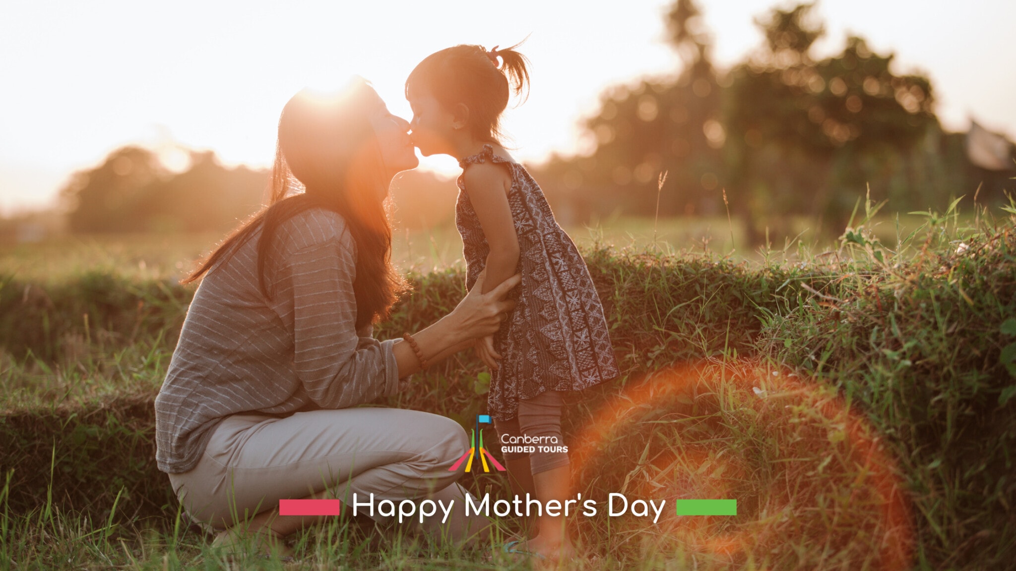 Mothers Day CGT 1 - Canberra Guided Tours A mother kneeling in a grassy field at sunset leans forward to kiss her young daughter, warm backlighting and lens flare highlighting them with a 'Happy Mother's Day' greeting and Canberra Guided Tours logo overlaid at the bottom.