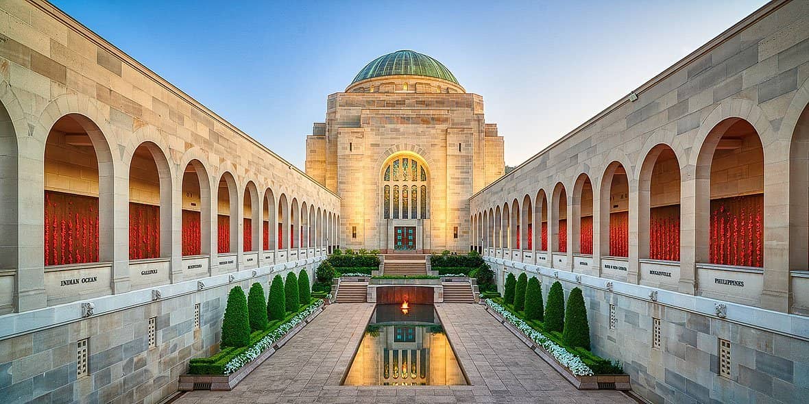calm contemplation australian war memorial canberra 9f9e75de 9483 45 To5NgjI 1 - Canberra Guided Tours Symmetrical courtyard of the Australian War Memorial at sunset, with a central reflecting pool and eternal flame leading to the domed memorial building flanked by colonnaded arcades displaying rows of red poppies.