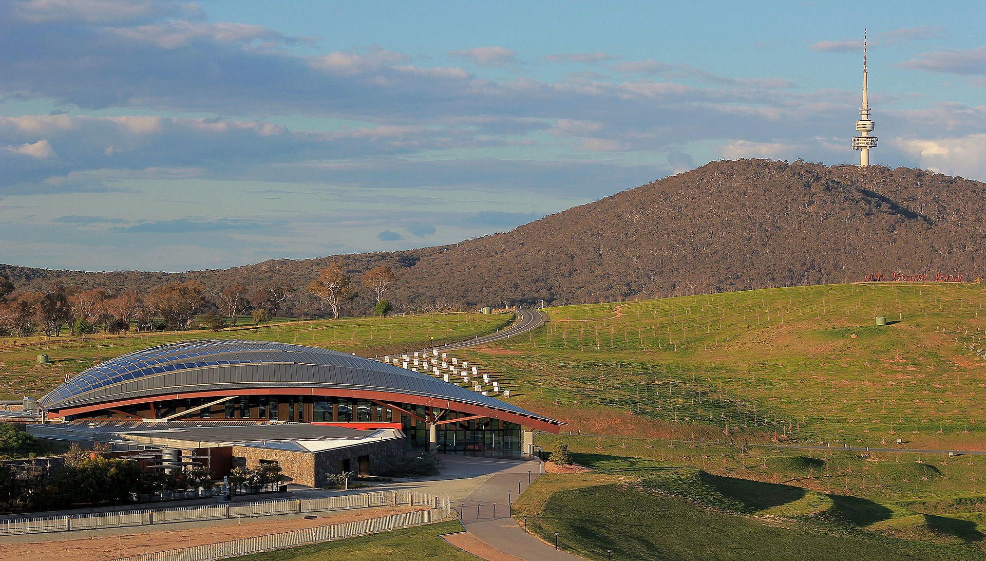 Highlights Tour National Arboretum - Canberra Guided Tours Curved glass-and-timber visitor centre set in green rolling hills with a winding road leading up to a tall communications tower on a tree-covered ridge under a partly cloudy sky.
