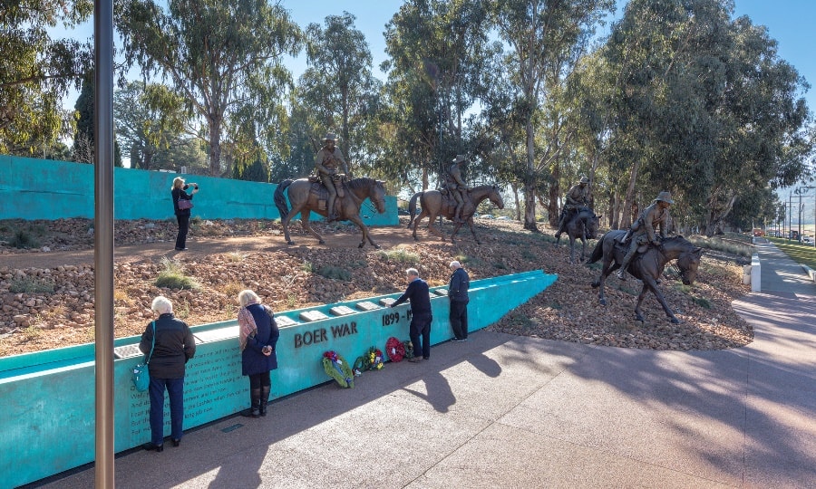IMG4 - Canberra Guided Tours Visitors pause at a turquoise memorial wall inscribed 'BOER WAR 1899–1902' with wreaths as bronze statues of mounted soldiers ride down a rocky slope amid tall eucalyptus trees.