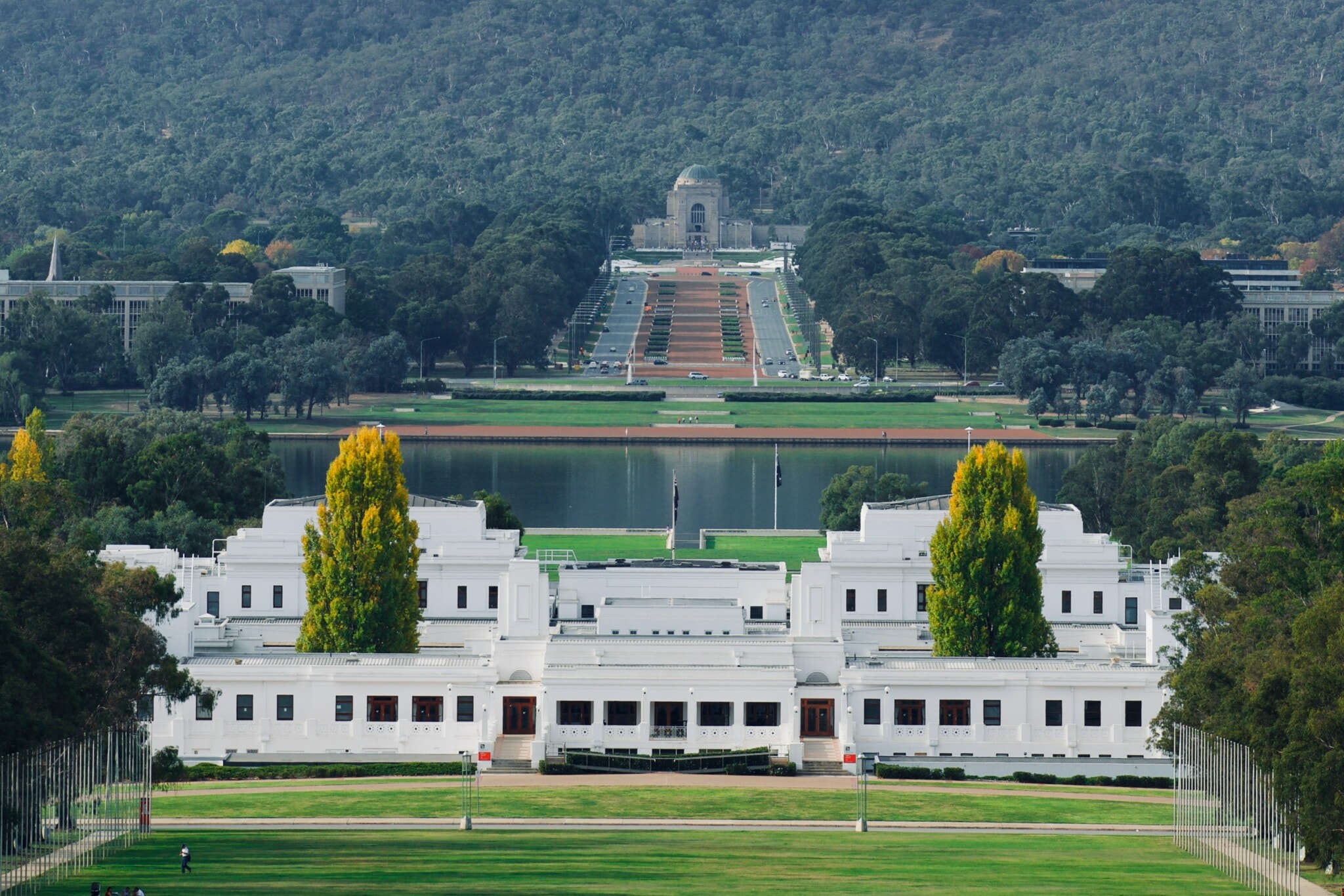 Provisional Parliament House - Canberra Guided Tours White neoclassical government building fronting a wide green lawn and reflecting lake, with a tree-lined ceremonial avenue leading uphill to a domed stone memorial against a densely wooded hillside.