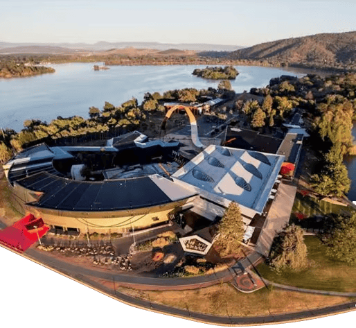 about us image - Canberra Guided Tours Aerial view of a curved waterfront complex featuring a dark-roofed circular building and an adjoining white canopy with oval skylights, an orange arched ramp, and surrounding trees by a calm lake with islands and distant hills.